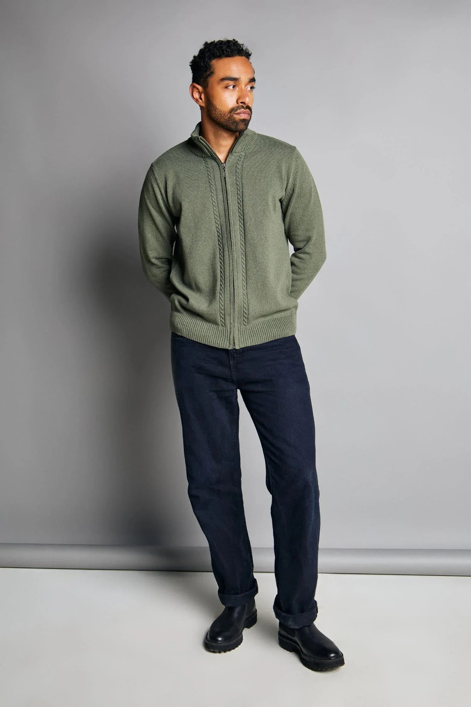 Hombre con chaqueta de punto verde, pantalón oscuro y botas negras sobre fondo gris